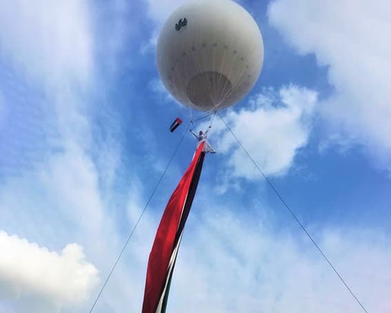 Helium balloon carrying an aerialist who unfurls a giant UAE flag against a blue daytime sky